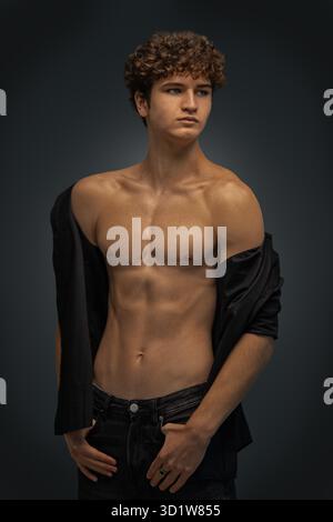 young man stands in a studio showcasing his curly hair and toned body. He wears an unbuttoned black shirt expressing a confident and relaxed demeanor during the photo shoot. Stock Photo