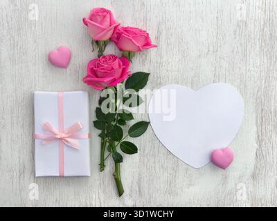 Pink flowers roses with paper card note in the shape heart with space for text, pink hearts on a white wooden background. Top view, flat lay Stock Photo
