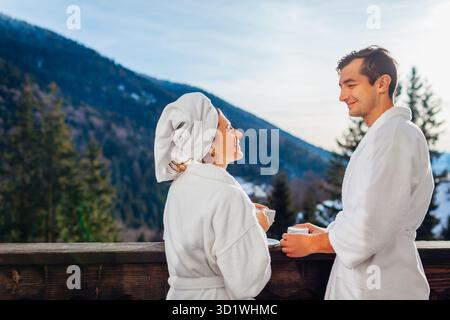 Young couple man and woman drinking coffee on hotel balcony enjoying winter mountains view wearing white robes during holidays. Vacation in the Carpat Stock Photo
