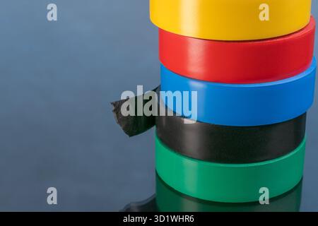 Stacked electrical tape rolls: yellow, red, blue, black, green on a reflective surface. Stock Photo