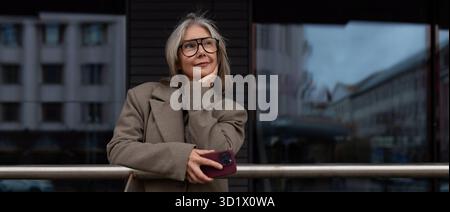 Middle-aged businesswoman stands thoughtfully next to a modern building, holding her phone in a business district on a cloudy da Stock Photo