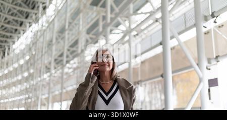 Middle-aged businesswoman in glasses talking on smartphone while walking through a modern airport terminal on a sunny day Stock Photo