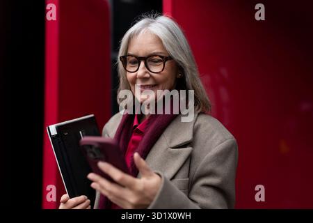 Elderly businesswoman checking her phone while holding a notebook outside in a bustling city during the afternoon Stock Photo
