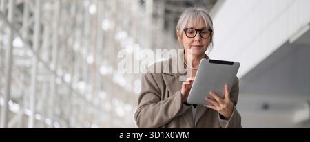 Middle-aged businesswoman using a tablet while standing in a modern office environment during daylight hours Stock Photo
