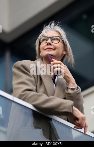 An older woman thoughtfully gazes into the distance while holding her phone on a modern balcony in an urban setting Stock Photo