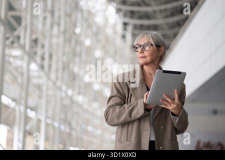 Stylish elderly woman with glasses using a tablet in a modern, well-lit architectural space during the afternoon Stock Photo