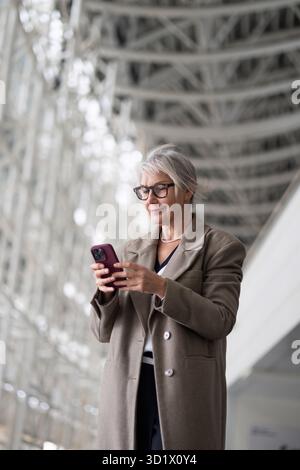 Stylish elderly Caucasian woman using her smartphone while standing indoors in a modern architectural space Stock Photo