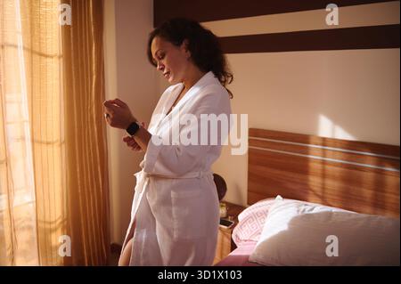 A woman in a white bathrobe stands by a sunlit bedroom window, adjusting a smartwatch. Warm light, wooden headboard, and soft pillows create an intima Stock Photo