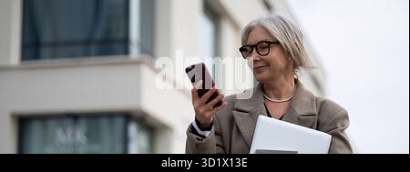 An older woman using a smartphone while carrying a laptop outside an office building on a cloudy day Stock Photo