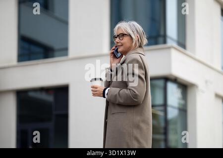 An older woman in a beige coat talking on her phone while holding a coffee cup outside a modern office building Stock Photo