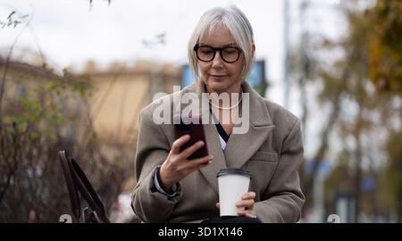 Stylish elderly woman enjoying a coffee while looking at her smartphone in an urban park during a cloudy afternoon Stock Photo