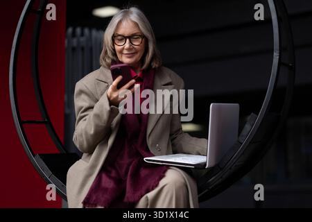 Senior businesswoman working outside on a laptop while checking her smartphone in a modern urban setting during the day Stock Photo