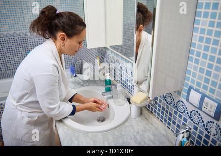 A woman in a white robe washes her hands at a marble sink, surrounded by blue mosaic tiles. The scene conveys cleanliness, personal care, and everyday Stock Photo