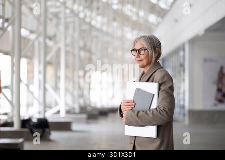 Older woman in a business suit holding a laptop and documents stands confidently in a modern office building during daytime Stock Photo