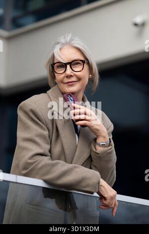 Middle-aged businesswoman stands confidently on a balcony, smiling, in a modern urban setting during the afternoon Stock Photo
