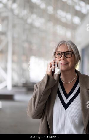 Senior businesswoman smiling and talking on her phone while walking outside in a modern urban setting during daylight Stock Photo