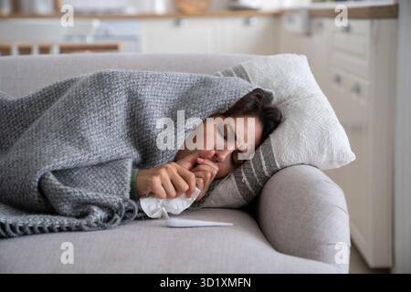 Sick unhealthy woman lying under blanket on sofa coughing and sneezing, using tissue to blow nose Stock Photo