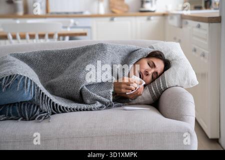 Sick unhealthy woman lying under blanket on sofa coughing and sneezing, using tissue to blow nose Stock Photo