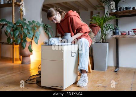 Creative woman sculptor making ceramic products sitting indoors in cozy workshop using potter wheel Stock Photo