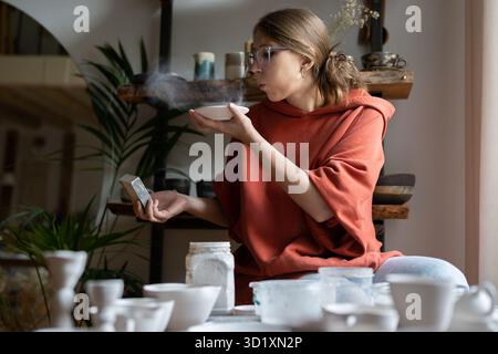 Woman potter using sandpaper, polishing pottery, sanding ceramics while working in cozy studio Stock Photo