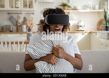 Surprised black woman with VR glasses sits on sofa clutch pillow in fear with astonished expression Stock Photo