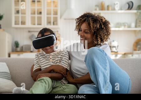 Happy joyful African American family mother and son using VR headset while relaxing together on sofa Stock Photo