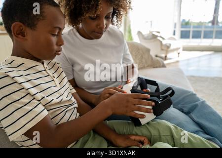 African American family mother and son sit on couch at home learning how to use VR glasses together Stock Photo