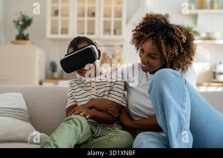 Happy joyful African American family mother and son using VR headset while relaxing together on sofa Stock Photo