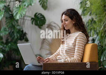 Freelancer young woman sitting in armchair in greenhouse, working on laptop surrounded by houseplant Stock Photo
