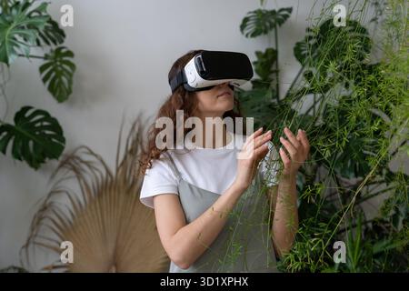Home gardening, metaverse. Woman using VR glasses in greenhouse, touching plant. Augmented reality. Stock Photo