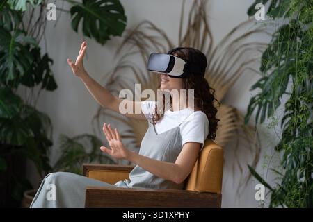Young woman wearing VR helmet experiencing virtual reality while resting in greenhouse Stock Photo