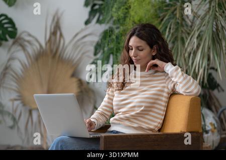 Pensive woman freelancer working on laptop in cozy garden office, female remote worker with computer Stock Photo