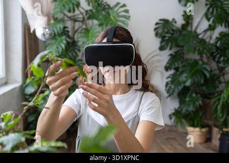 Home gardening, metaverse. Woman using VR glasses in greenhouse, touching plant. Augmented reality. Stock Photo
