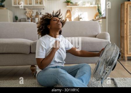 Young African American woman cooling down by ventilator at home, feeling unwell during heatwave Stock Photo