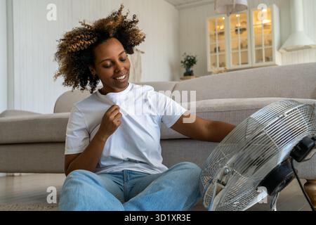 Pleased joyful black woman sit on floor cools holds ventilator regulates airflow at home in heat day Stock Photo
