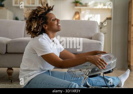 Happy young black woman sits in front of fan at home cooling down at home after being in heat Stock Photo
