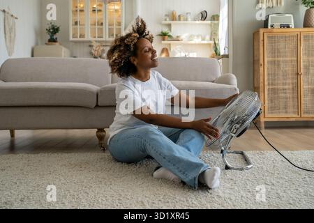 Satisfied happy young African American woman using fan at home during extreme summer heat Stock Photo