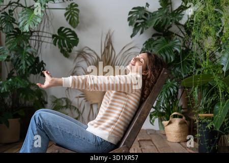 Serene relaxed young woman with closed eyes stretching arms, relaxing in urban jungle interior Stock Photo