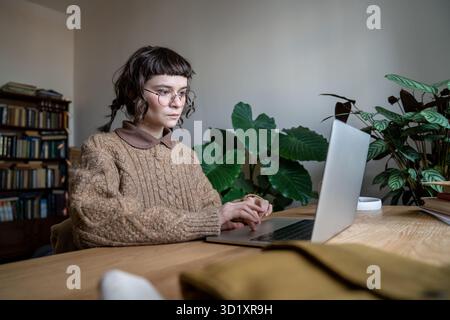 Focused teen student girl wearing glasses sitting at table typing on laptop Stock Photo