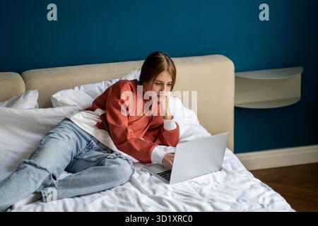 Thoughtful interested teen girl student lying on bed use laptop college study online webinar at home Stock Photo
