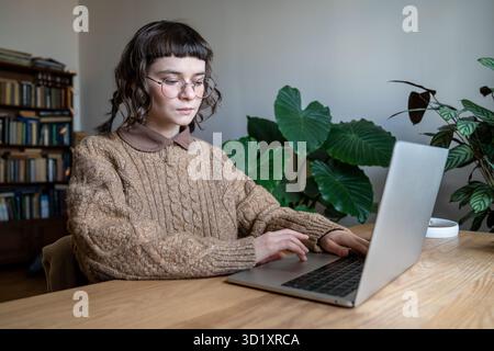 Serious girl student in eyeglasses sitting at table with laptop. Aesthetically place for home office Stock Photo
