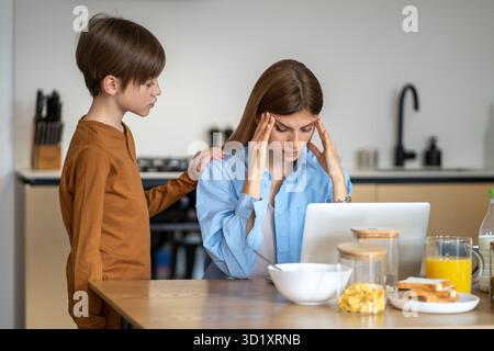 Worried child boy touches mother sitting at kitchen table with laptop from fatigue holding temples Stock Photo