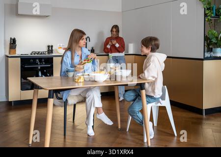 Family breakfast on kitchen, mom son sit at table eat, daughter chatting in phone in social media. Stock Photo