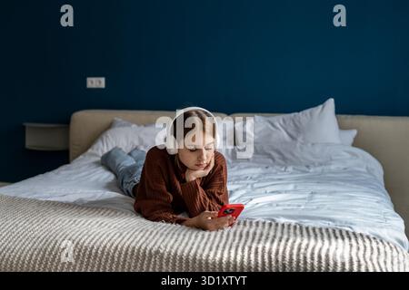 Tranquil teen girl in headphones relaxed lies on bed using phone surfing social media posts at home Stock Photo