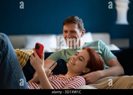 Happy smiling man and woman lying relaxed on sofa look screen phone. Positive family online shopping Stock Photo