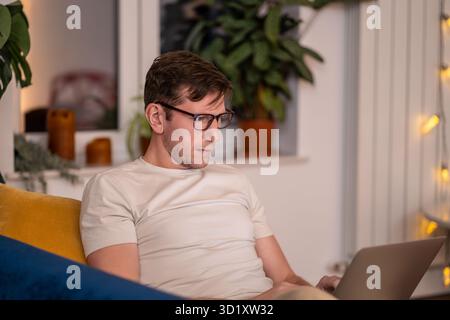 Man sits on couch with laptop on lap works overtime. Remote online internet studying work from home Stock Photo