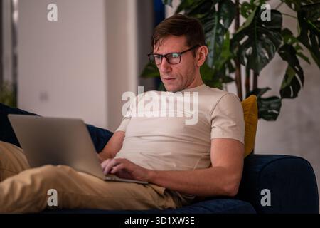 Man middle aged student working on educational project lying on couch at home in evening after job. Stock Photo