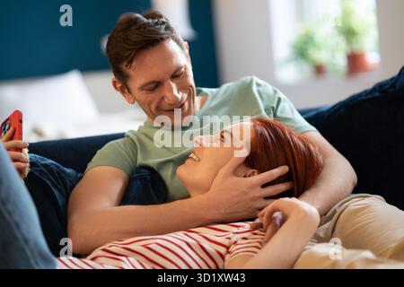 Happy family couple lovingly look at each other relaxing lying on sofa together sharing care at home Stock Photo