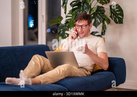 Smiling positive man freelancer talks phone works on laptop sits on couch at home in evening. Stock Photo