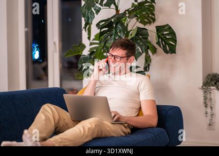Laughing man in eyeglasses sitting on sofa with laptop on knees having fun cellphone conversation Stock Photo